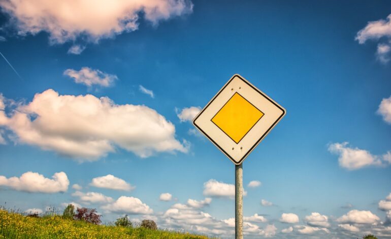 Ein Verkehrsschild, das Vorfahrtsrecht zeigt. Dahinter Blauer Himmel mit Wolken und grüne Wiese.