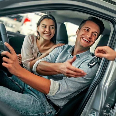 Ein Fahrer und eine Beifahrerin sitzen in einem neuen Auto. Dem Fahrer wird der Autoschlüssel überreicht.