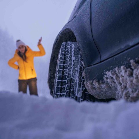 Auto im Schnee im Vordergrund, Fokus von unten auf Vorderreifen, Schnee hängt am Unterboden. Frau in Winterbekleidung im Hintergrund hebt ratlos eine Hand.