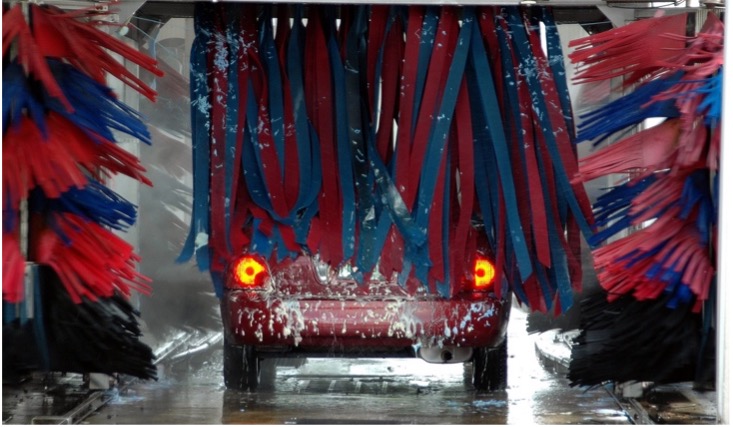 Rotes Auto mit leuchtenden Rücklichtern zwischen den Bürsten der laufenden Waschanlage.