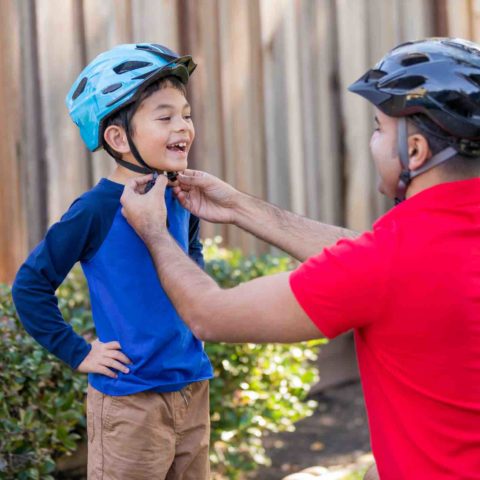 Mann mit Fahrradhelm und rotem Shirt kniet vor kleinem Jungen mit blauem Shirt und setzt ihm den Helm auf.