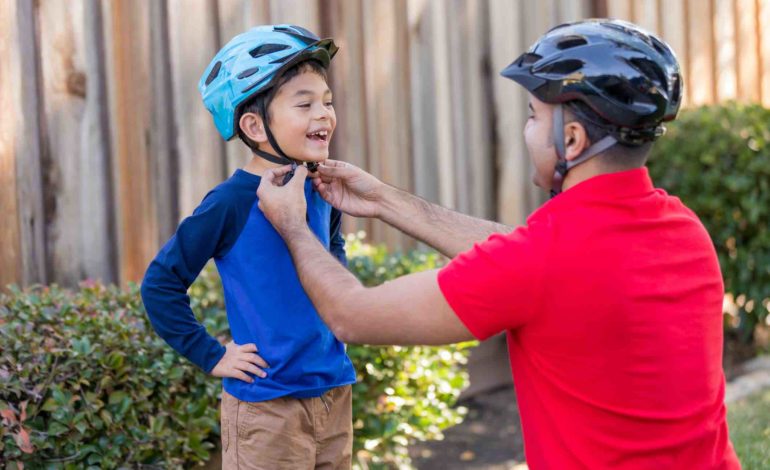 Mann mit Fahrradhelm und rotem Shirt kniet vor kleinem Jungen mit blauem Shirt und setzt ihm den Helm auf.