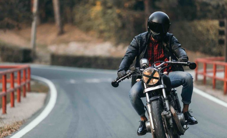 Motorradfahrer mit schwarzem Helm, Lederjacke und Jeans fährt auf einer Straße auf die Kamera zu.