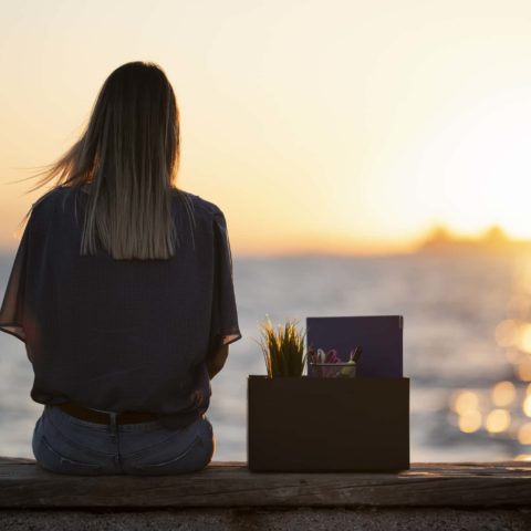 Junge Frau sitzt mit dem Rücken zur Kamera auf einer Mauer. Neben ihr steht eine Box mit Unterlagen, Schreibutensilien und einer Pflanze. Im Hintergrund ist ein Sonnenuntergang über dem Meer zu sehen.