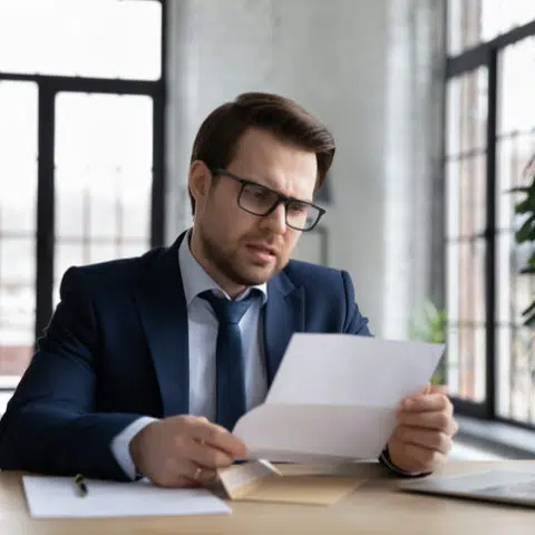 Mann mit Anzug, Krawatte und Brille liest mit besorgtem Gesichtsausdruck einen Brief am Schreibtisch. Auf dem Schreibtisch steht ein Laptop, im Hintergrund sind hohe Fenster.