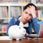 Student beantragt BAföG am Laptop, fasst sich dabei an den Kopf. Im Vordergrund ein weißes Sparschwein, im Hintergrund ein Bücherregal.