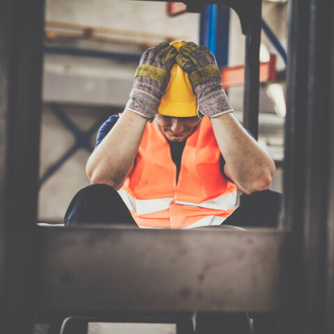 Ein Bauarbeiter mit orangefarbener Weste, gelbem Helm und Handschuhen sitzt auf einer Baustelle und hält sich frustriert die Hände an den Kopf, wegen Schadensersatz bei Bauverzug.