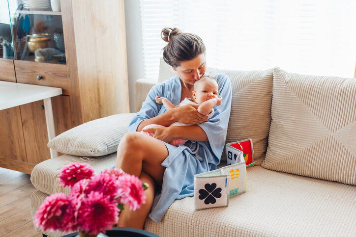 Mutter während der Elternzeit mit Baby und Babybuch auf dem Sofa. Davor eine Vase mit Blumen, dahinter die Küche und ein Tisch.