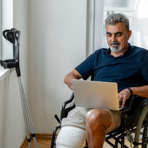 Mann im Rollstuhl nach Unfall in der Mittagspause. Auf dem Schoß hat er einen Laptop, am Fenster daneben lehnen Krücken.