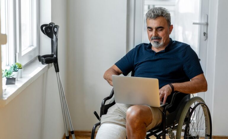 Mann im Rollstuhl nach Unfall in der Mittagspause. Auf dem Schoß hat er einen Laptop, am Fenster daneben lehnen Krücken.
