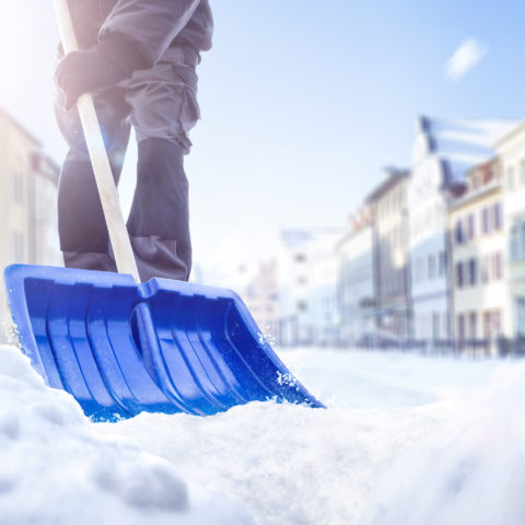 Eine Person benutzt eine Schneeschaufel auf einer verschneiten Straße. Im Hintergrund sieht man einige Häuser.