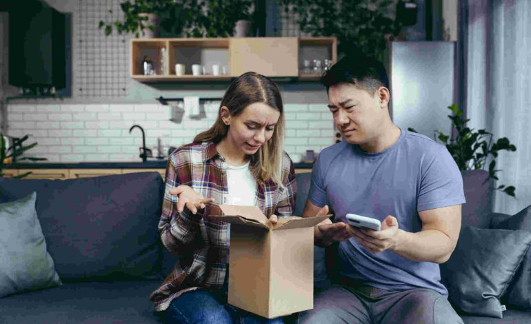 Eine junge Frau und ein junger Mann sitzen auf dem Sofa, ein geöffnetes Paket auf dem Schoß. Beide sehen enttäuscht aus. Der Mann hält ein Smartphone in der Hand. Im Hintergrund ist eine Küche zu sehen.