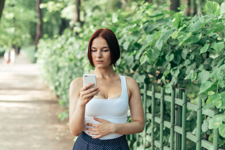 Eine junge Frau auf einem Waldweg, im Hintergrund ein Zaun und eine Hecke. Sie blickt auf ihr Smartphone und prüft ihre Erreichbarkeit in der Freizeit.