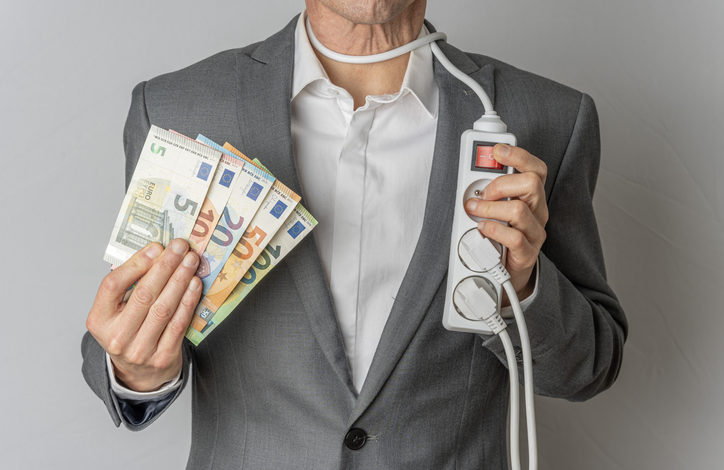 Ein Mann im Anzug hält in einer Hand mehrere Euro-Scheine, in der anderen eine Mehrfachsteckdose. Das Kabel ist um seinen Hals gewickelt.