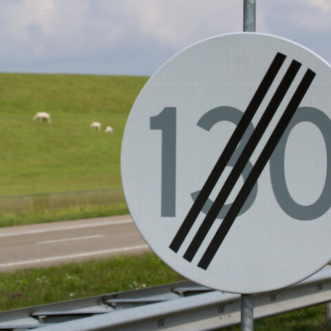 Schild mit Geschwindigkeitsbegrenzung 130 durchgestrichen in Ländern ohne Tempolimit, im Hintergrund eine Straße und ein Hügel mit Schafen.