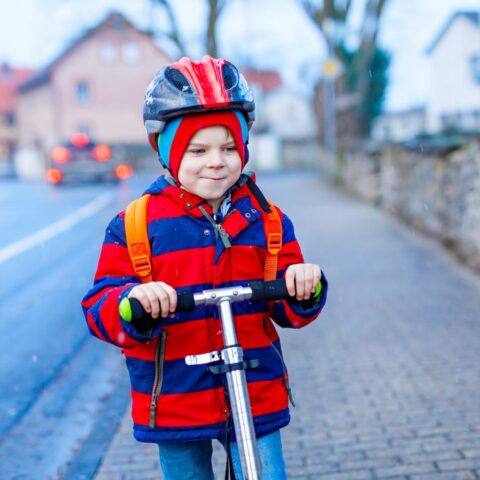 Ein Kind im Straßenverkehr auf einem Roller, mit Helm und Jacke.