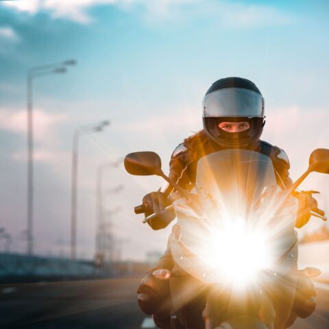 Ein Motorradfahrer mit Schutzkleidung fährt auf der Straße auf die Kamera zu, das Licht blendet. Dahinter hohe Straßenlaternen.