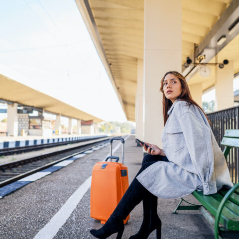 Frau sitzt am Bahnsteig, prüft mit dem Smartphone ihre Rechte bei Zugverspätung, neben sich einen orangefarbenen Koffer.