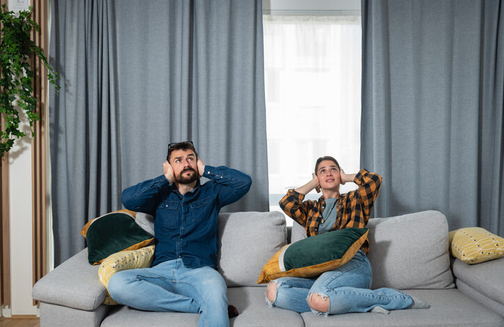 Ein Mann und eine Frau an Sonn- und Feiertagen auf einem Sofa vor einem Fenster mit Vorhängen. Beide halten sich die Ohren zu und blicken nach oben, wegen der Lärmbelästigung.