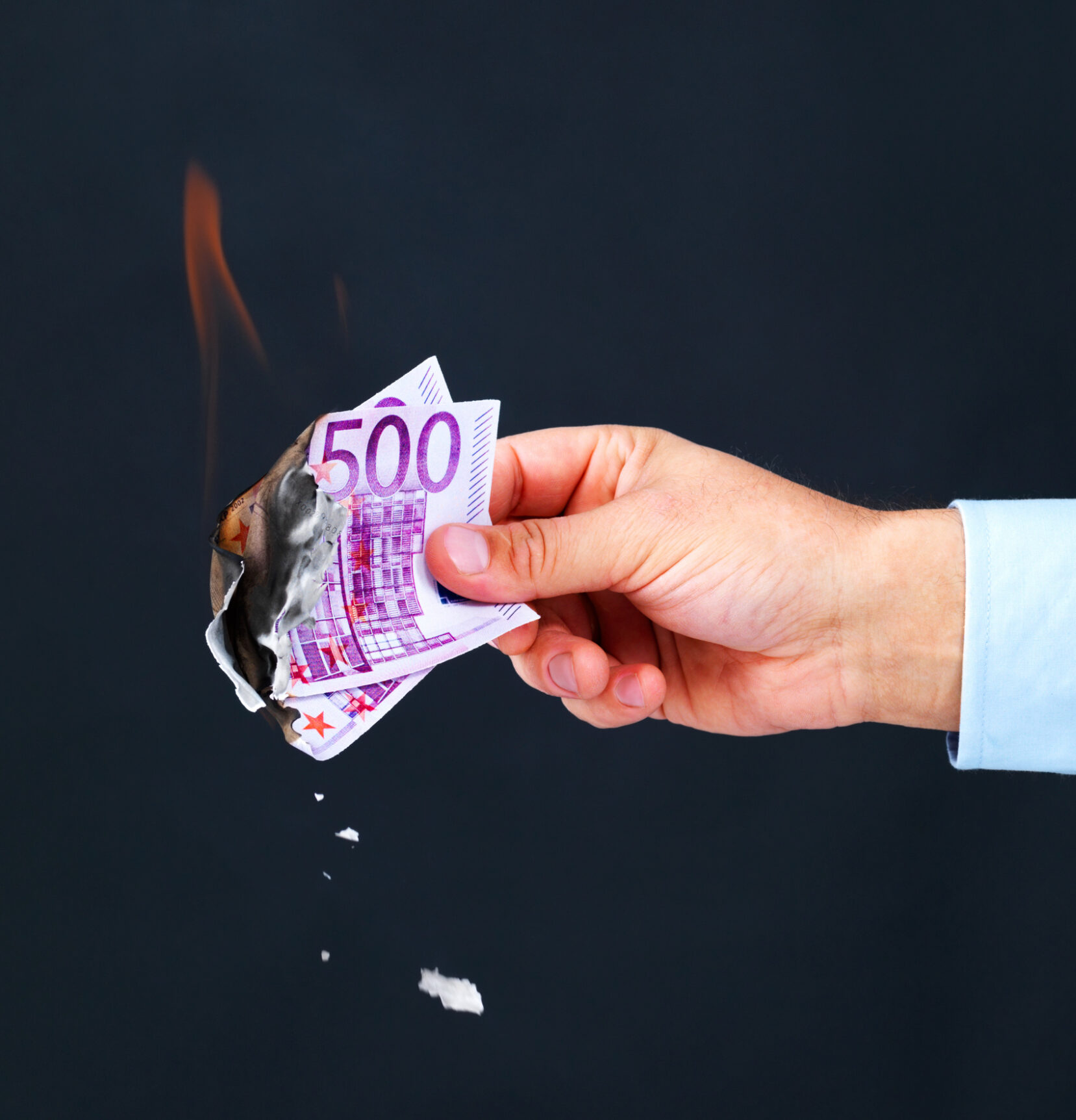 Beschädigter Geldschein umtauschen:Eine Hand hält 500-Euro-Scheine fest, die brennen, vor einem schwarzen Hintergrund.
