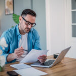 Schuldnerverzeichnis: Mann mit Brille sitzt vor einem Laptop, mit Stift und Papier in den Händen.