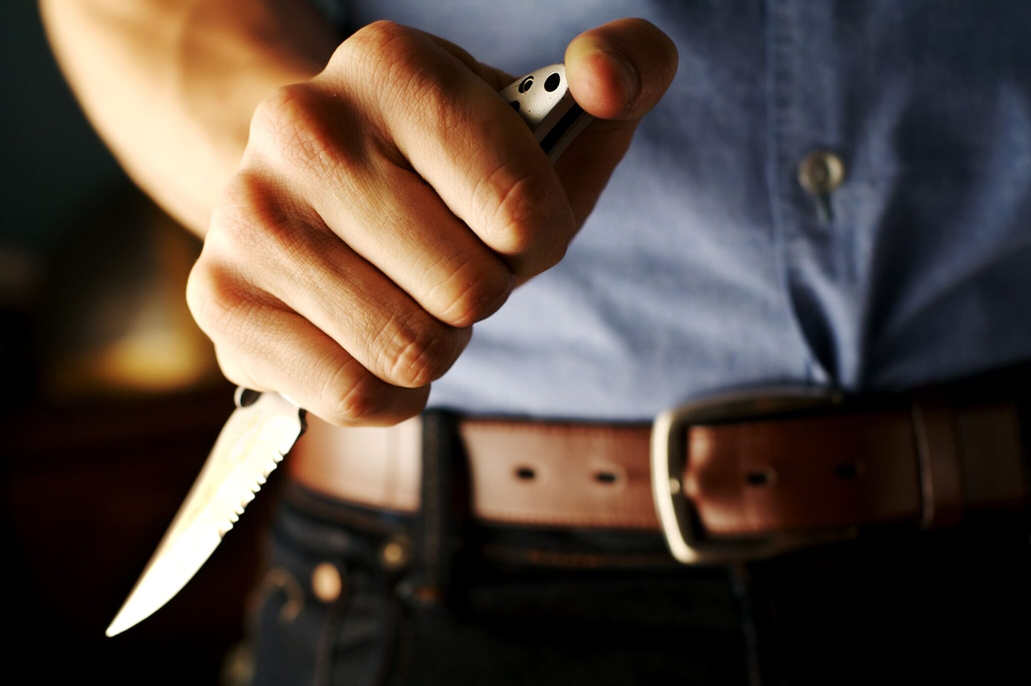 Eine Männerhand, die ein kurzes Messer hält. Dahinter sind Hemd, Gürtel und Hose des Mannes zu sehen.
