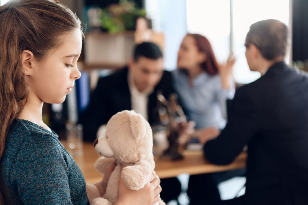 Mädchen mit Teddybär, im Hintergrund Eltern, die sich darüber streiten, wie lang man Unterhalt zahlen muss.