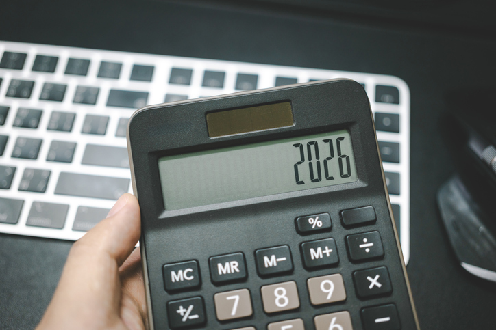 Ein Taschenrechner in einer Hand zeigt die Zahl 2026, dahinter ist eine Computer-Tastatur.