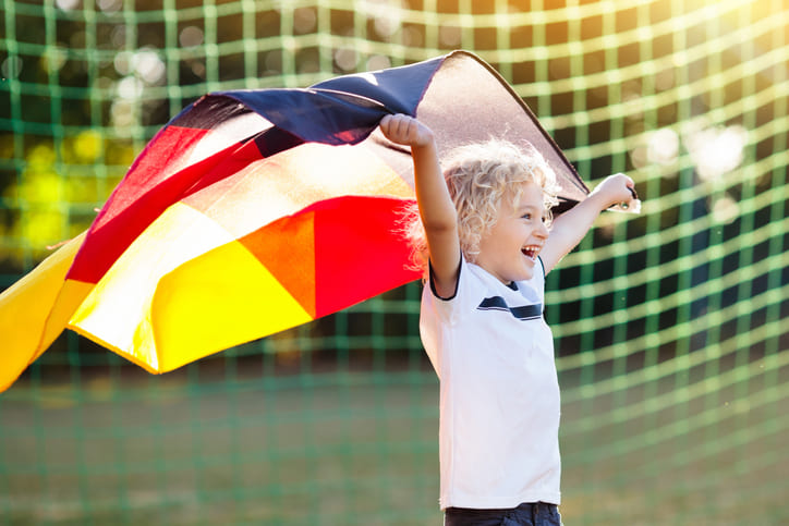 Ein Kind im Deutschland Trikot steht vor einem Fußballtor und hält eine Deutschlandflagge hoch.