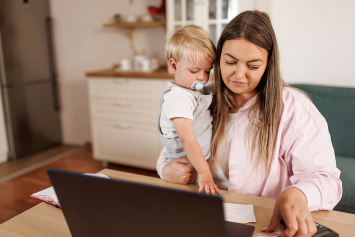 Frau sitzt am Laptop zum Elternzeit beantragen, ihr Kind mit Schnuller auf dem Schoß.