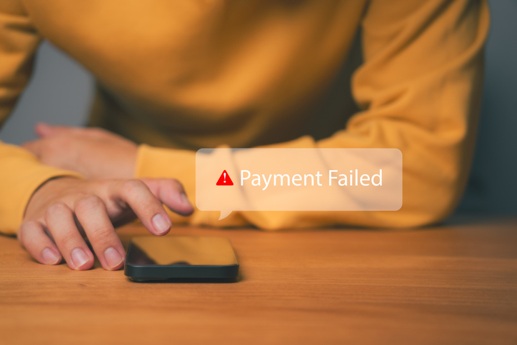Oberkörper und Hände einer Frau mit gelbem Oberteil, die auf einem Smartphone tippt, das auf einem Holztisch liegt. Darüber steht mit einem roten Ausrufezeichen Payment Failed.
