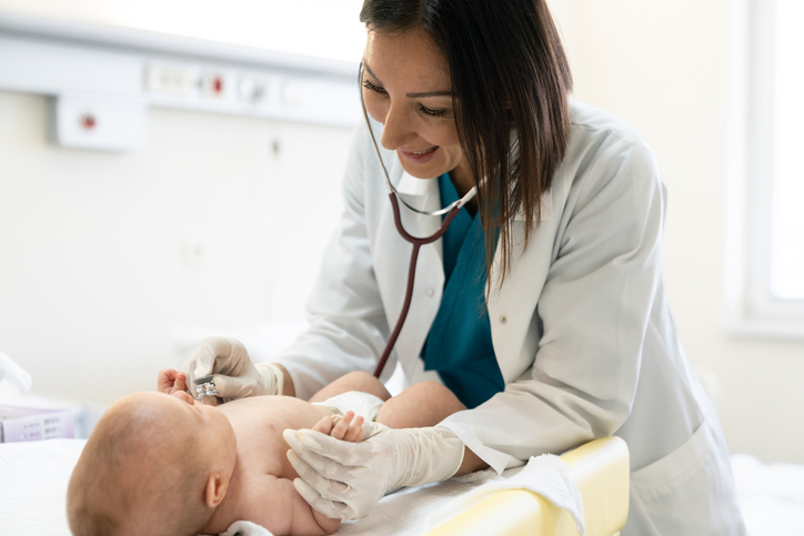 Ärztin mit Stethoskop um den Hals beugt sich über ein Baby und hält dessen Finger fest, sie führt die Neuerungen ab Mai 2026 im Neugeborenen-Screening durch.