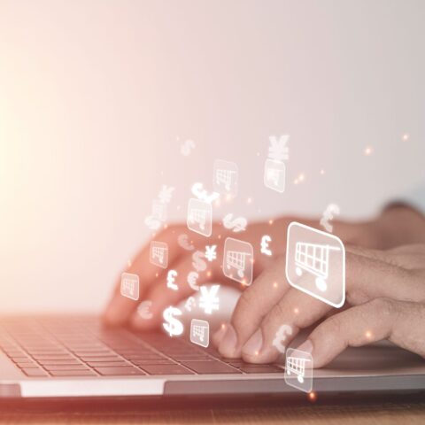 Zwei Hände tippen auf der Tastatur eines Laptops, suchen nach China fake Shops. Darüber Symbole von Einkaufswägen und verschiedenen Währungen.