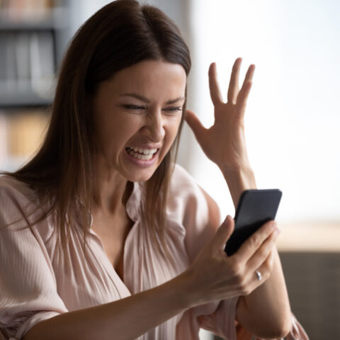 Junge Frau hebt eine Hand und ärgert sich über unerwünschte Werbung auf dem Smartphone in ihrer anderen Hand.