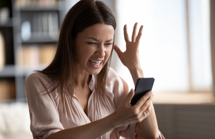 Junge Frau hebt eine Hand und ärgert sich über unerwünschte Werbung auf dem Smartphone in ihrer anderen Hand.