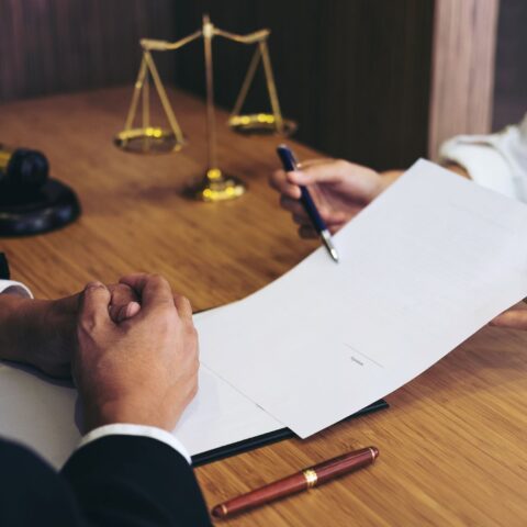 Die Hände zweier Personen am Tisch. Eine Person hält der anderen ein Papier und einen Stift zum Unterschreiben der Erstattung der außergerichtlichen Anwaltskosten hin. Dahinter ein Richterhammer und eine goldene Waage.